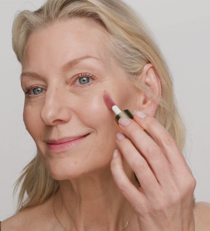 Model applying Vibrant Cheeks Liquid Blush in Dusty Rose on cheeks using a makeup brush for a natural glow.